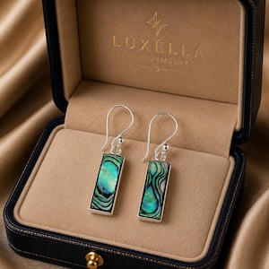May include: A pair of silver drop earrings featuring rectangular inlays of iridescent green and blue abalone shell. The earrings are displayed inside a navy blue jewelry box with the gold text LUXELLA FINE JEWELRY printed on the interior lid.