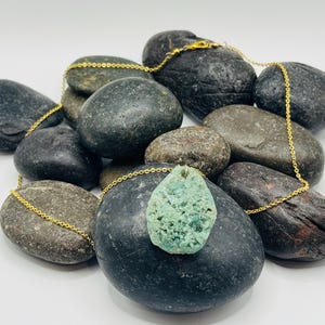 Può includere: Una collana con catena dorata e un ciondolo in pietra verde. Il ciondolo ha una forma irregolare e ruvida, con un aspetto naturale e organico. La collana è esposta su un letto di pietre lisce e grigie.