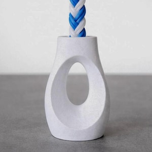 Könnte beinhalten: Ein weißer, strukturierter Lichtzug aus Keramik mit einem ovalen Ausschnitt. Ein geflochtenes Kabel in Weiß und Blau erstreckt sich von oben. Der Lichtzug hat ein glattes, geschwungenes Design und ein mattes Finish. Der Hintergrund ist ein neutrales Grau.