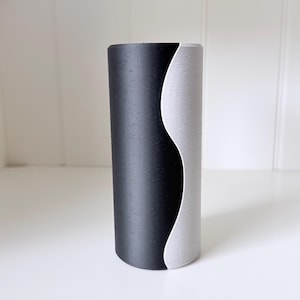 Può includere: Un vaso cilindrico bicolore. Una metà è nera, l'altra grigio chiaro, separate da una linea curva. Il vaso ha una finitura strutturata ed è esposto su una superficie bianca. Misura circa 20 cm di altezza.