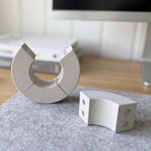 May include: A light gray, segmented desk organizer in a curved shape, with a matching small block. The organizer is made of a textured material and sits on a gray felt mat. The design is modern and minimalist.