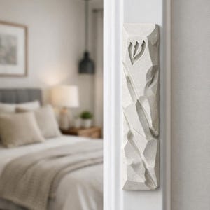 May include: A textured, off-white mezuzah case with a carved design and a Hebrew letter. The rectangular case is mounted on a white door frame, with a blurred bedroom scene in the background. The mezuzah is a religious item.