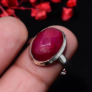 Puede incluir: Un anillo de plata con una gran piedra preciosa de color rojo intenso, forma ovalada y tallada en facetas, engastada en un bisel de metal liso. La piedra muestra una superficie texturizada y reflectante, con una banda de metal plateado pulido.