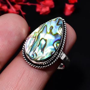 May include: A teardrop-shaped abalone shell ring set in a silver band with a twisted rope-style border. The iridescent shell displays swirling patterns of green, blue, and purple hues, securely held within a polished metal setting.
