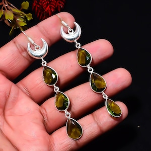May include: A pair of silver dangle earrings featuring a crescent moon top and three faceted, pear-shaped olive green gemstones suspended in a vertical line. The earrings have a polished silver finish and secure with a hook-style closure.