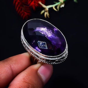 May include: A sterling silver ring featuring a large, oval-shaped, faceted deep purple amethyst gemstone. The silver setting displays intricate beaded and filigree metalwork along the edges, surrounding the central stone in a secure bezel mount.