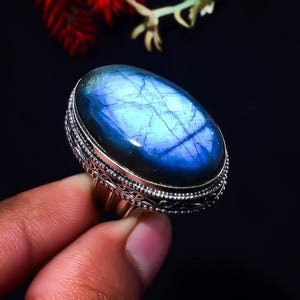 May include: An oval-shaped labradorite gemstone ring featuring a prominent blue iridescent flash across its surface. The stone is set in a polished silver band with intricate, decorative metalwork detailing along the outer edge of the setting.