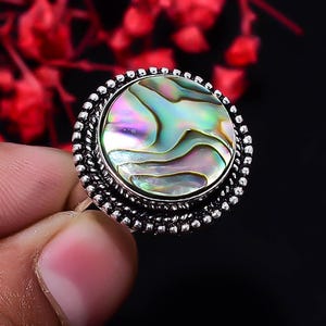 Puede incluir: Un anillo redondo con una incrustación de concha de abulón pulida e iridiscente con tonos arremolinados en verde, morado y azul. La piedra central está engastada en un marco de plata oxidada texturizada con borde de cuentas y detalle trenzado.