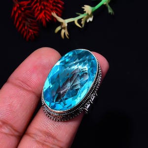 May include: A ring featuring a large, oval-shaped, faceted sky-blue gemstone set in a decorative silver band. The metal setting displays intricate beaded and textured detailing around the perimeter of the vibrant, translucent blue stone.