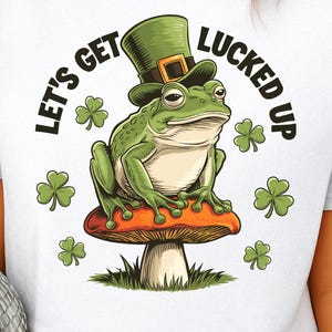 Puede incluir: Camiseta blanca con una rana verde con un sombrero de duende verde, sentada en una seta roja y blanca. El texto "LET'S GET LUCKED UP" está arqueado sobre la rana, con tréboles verdes esparcidos.