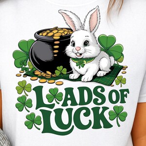 May include: White t-shirt featuring a cartoon rabbit with a green shamrock collar, sitting next to a pot of gold. The words "Loads of Luck" are printed in green with shamrock accents. A festive design for St. Patrick's Day.