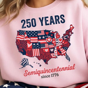 Puede incluir: Sudadera rosa claro con el texto "250 YEARS" sobre un mapa de los Estados Unidos decorado con diseños patrióticos. El texto "Semiquincentennial since 1776" está debajo. Parte de una colección de ropa patriótica.