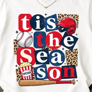 Puede incluir: Camiseta blanca con un diseño temático de béisbol. El diseño presenta el texto "Tis the Season" en bloques rojos y azules, junto con una pelota de béisbol, un casco, palomitas de maíz y un bate.