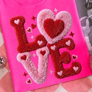 Puede incluir: Una sudadera rosa brillante con la palabra "LOVE" bordada en hilo de lana roja y rosa, con detalles de corazones y estrellas. En el fondo, una revista con el titular "Better Late Than".