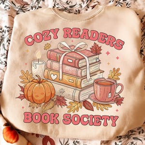Puede incluir: Una sudadera beige de cuello redondo con un estampado gráfico de una pila de tres libros atados con una cinta blanca, una calabaza, una vela encendida y una taza de té. El texto COZY READERS BOOK SOCIETY aparece en letras de color rosa.