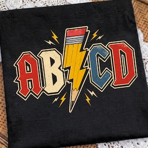 Puede incluir: Una camiseta negra con un estampado gráfico de las letras AB y CD en una fuente roja y azul desgastada. Entre las letras hay un lápiz amarillo con forma de rayo y goma rosa, rodeado de pequeños rayos amarillos.
