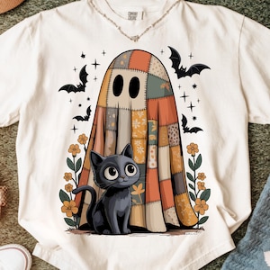 Puede incluir: Una camiseta color hueso con un gráfico de un fantasma cubierto por una colcha de retazos detrás de un gato negro. El diseño incluye flores naranjas a ambos lados, murciélagos negros y pequeñas estrellas dispersas alrededor de las figuras.