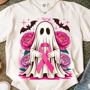 Puede incluir: Una camiseta color hueso con un gráfico de un fantasma blanco sosteniendo un lazo de concienciación rosa. El diseño incluye peonías rosas vibrantes, dos murciélagos negros y pequeños detalles de estrellas rosas. La camiseta tiene cuello redondo.