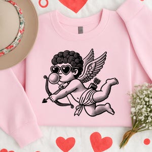 Puede incluir: Sudadera rosa claro con una ilustración en blanco y negro de Cupido con gafas de sol en forma de corazón, soplando una burbuja y sosteniendo un arco y una flecha. El diseño incluye alas y un carcaj de flechas. La sudadera es de color rosa liso.