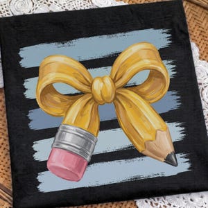 Puede incluir: Un cuadrado de tela negro con un estampado gráfico de un lápiz amarillo con forma de lazo. El lápiz tiene una virola plateada metálica y una goma de borrar rosa en un extremo, con una punta de grafito afilada en el otro, sobre trazos azul claro.