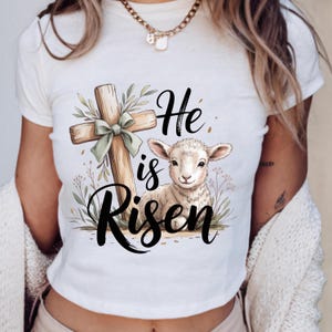 Puede incluir: Camiseta blanca con un diseño gráfico. El diseño incluye una cruz de madera con un lazo verde, un cordero y las palabras "He is Risen" en escritura negra, con elementos florales.