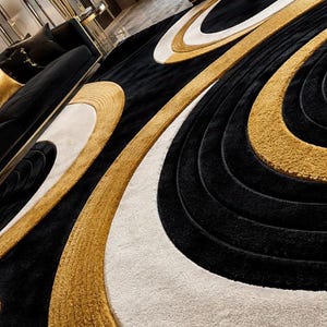 Pode incluir: Um tapete grande com um padr&atilde;o repetitivo de formas ovais conc&ecirc;ntricas em preto, branco e dourado. O design consiste em faixas texturizadas e elevadas de pelo dourado e preto que se alternam com sec&ccedil;&otilde;es brancas lisas e uniformes.