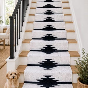 May include: A white stair runner with a repeating black geometric diamond pattern installed on wooden stairs. The runner features a textured weave and a solid tan border at the base of the bottom step, covering the center of each individual stair tread.