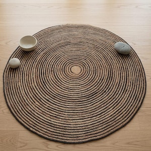 May include: A round rug featuring a concentric circular pattern in alternating shades of tan and dark brown. The rug is textured with a coiled rope-like construction. Two smooth, light-coloured stones and a small white bowl are placed on the rug.