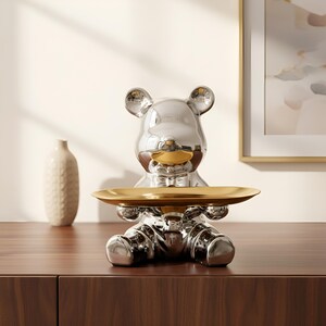 Puede incluir: Una bandeja decorativa plateada con forma de oso con una bandeja dorada sostenida en sus brazos. El oso tiene una superficie brillante y reflectante y una nariz dorada. La bandeja está sobre una superficie de madera marrón oscuro.