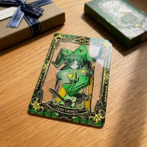 Puede incluir: Una tarjeta acrílica transparente con una ilustración verde y amarilla de un personaje con un traje de bufón. La tarjeta está enmarcada en un borde verde decorativo con las palabras "JADE CROWN" y "UP-001". Una caja de regalo con una cinta azul está en el fondo.