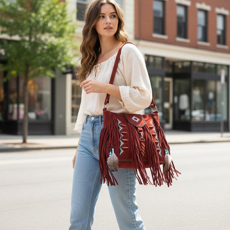 May include: A burgundy suede shoulder bag with long fringe and decorative beading. The bag features a geometric design on the flap and is adorned with feathers. The bag is carried over the shoulder.