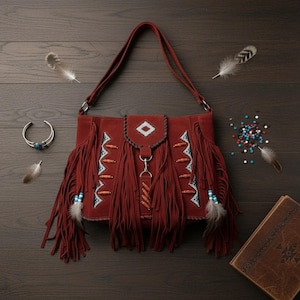 May include: A rust-colored suede shoulder bag with long fringe and decorative beading. The bag features a flap with a geometric design and a long strap. A silver bracelet with turquoise stones and feathers are nearby.