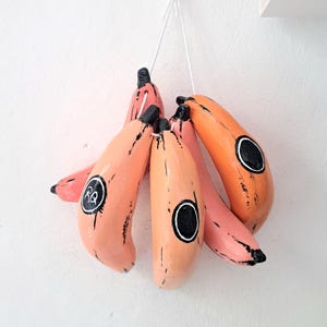 May include: A cluster of six painted banana-shaped ornaments in shades of pink and orange, hanging against a white wall. Each banana has a black tip and a black circle detail. One banana has the letters "RQ" in a circle.
