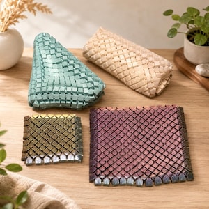 May include: Four square-shaped tile mats featuring a grid of small, raised, interlocking geometric pieces. One mat is light blue, one is beige, one is a small gold-toned metallic, and one is a larger purple-toned metallic with a dark border.