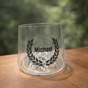Könnte beinhalten: Ein klares Glas mit einem schwarzen Lorbeerkranz-Design und dem Namen "Michael" in der Mitte. Das Glas steht auf einer Holzoberfläche mit verschwommenem grünem Hintergrund.