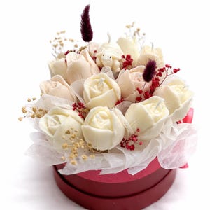 Puede incluir: Una caja de regalo redonda, color burdeos, llena de velas con forma de rosa blancas y crema, acentuadas con pequeñas bayas rojas, flores secas y una pequeña figura de tortuga. Las velas están dispuestas sobre papel de seda blanco.