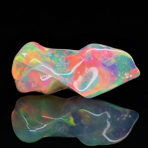 May include: A polished opal gemstone with a distinctive, flowing form. The opal exhibits a vibrant play of colour, including shades of pink, orange, green, and blue. The gemstone is set against a black background, with its reflection visible below.