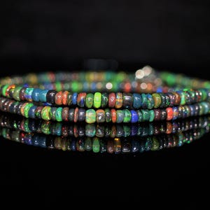 May include: A strand of small, circular, multi-colored opal beads featuring a dark base with vibrant flashes of green, blue, orange, and red. The beads are arranged in a continuous loop, displaying a polished texture and reflective surface.