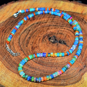 May include: A beaded necklace featuring small, round, multi-colored opal beads in shades of blue, orange, green, and pink. The necklace includes a silver-toned metal lobster claw clasp and an adjustable chain link extender at the end of the strand.