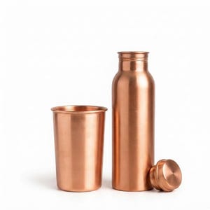 Puede incluir: Una botella de agua de cobre pulido con un vaso cilíndrico a juego y una tapa de rosca separada. La botella presenta un acabado metálico liso con un cuello estrecho y una base ancha, mientras que el vaso tiene un diseño sencillo de paredes rectas.