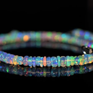 May include: A beaded bracelet composed of iridescent opal gemstones. The bracelet features a variety of colors, including blue, green, orange, and pink. The beads are small and round, strung together to form a flexible band. The bracelet is displayed on a reflective black surface.