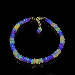 May include: A bracelet composed of small, round, iridescent opal beads featuring a vibrant color palette of blue, purple, and green hues. The jewelry piece includes a gold-toned metal chain extender with a lobster clasp closure mechanism.