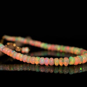 May include: A colorful opal bead necklace with a gold clasp. The beads are a mix of orange, pink, green, and yellow, and are arranged in a strand. The necklace is displayed on a reflective black surface.