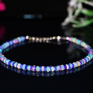 May include: A delicate bracelet composed of small, colorful opal beads. The beads display a range of blues, pinks, and greens, strung together with a silver clasp. The bracelet is displayed on a reflective black surface.