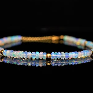Pode incluir: Uma pulseira delicada com contas de opala iridescentes em tons de azul, branco e toques de rosa. As contas são unidas por elos de metal dourado e pequenas contas douradas. A pulseira é exibida em uma superfície preta reflexiva.