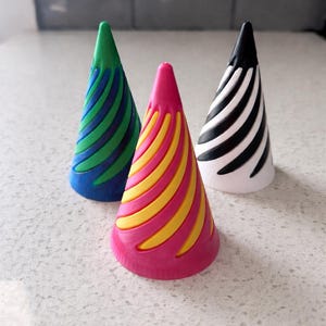 May include: Three colorful party hats. One is green and blue, another is pink and yellow, and the last is black and white. The hats have a spiral design and are arranged on a white surface.