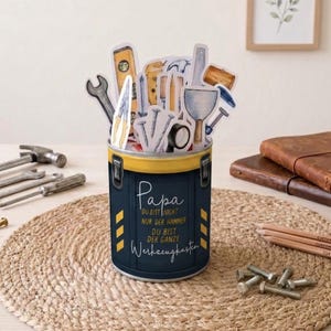 May include: A cylindrical container with a yellow rim and a dark blue body, decorated with the text "Papa" and "Werkzeugkasten". The container is filled with tool-shaped cutouts, including a level, wrench, and hammer. A woven mat and pencils are in the background.
