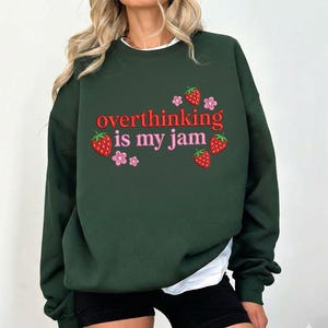 May include: Dark green sweatshirt with the phrase "overthinking is my jam" in red and pink lettering. The design includes strawberry and flower graphics. The sweatshirt is a casual style.