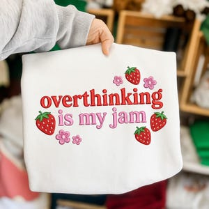 May include: A white sweatshirt with the embroidered phrase "overthinking is my jam" in red and pink. The design includes red strawberries and pink flowers, creating a playful and whimsical aesthetic.