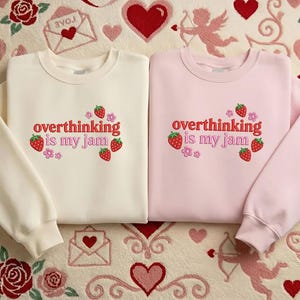 May include: Two crewneck sweatshirts, one cream and one pink, with the phrase "overthinking is my jam" embroidered in red. The design includes strawberries and flowers. The sweatshirts are displayed on a patterned surface with hearts and roses.